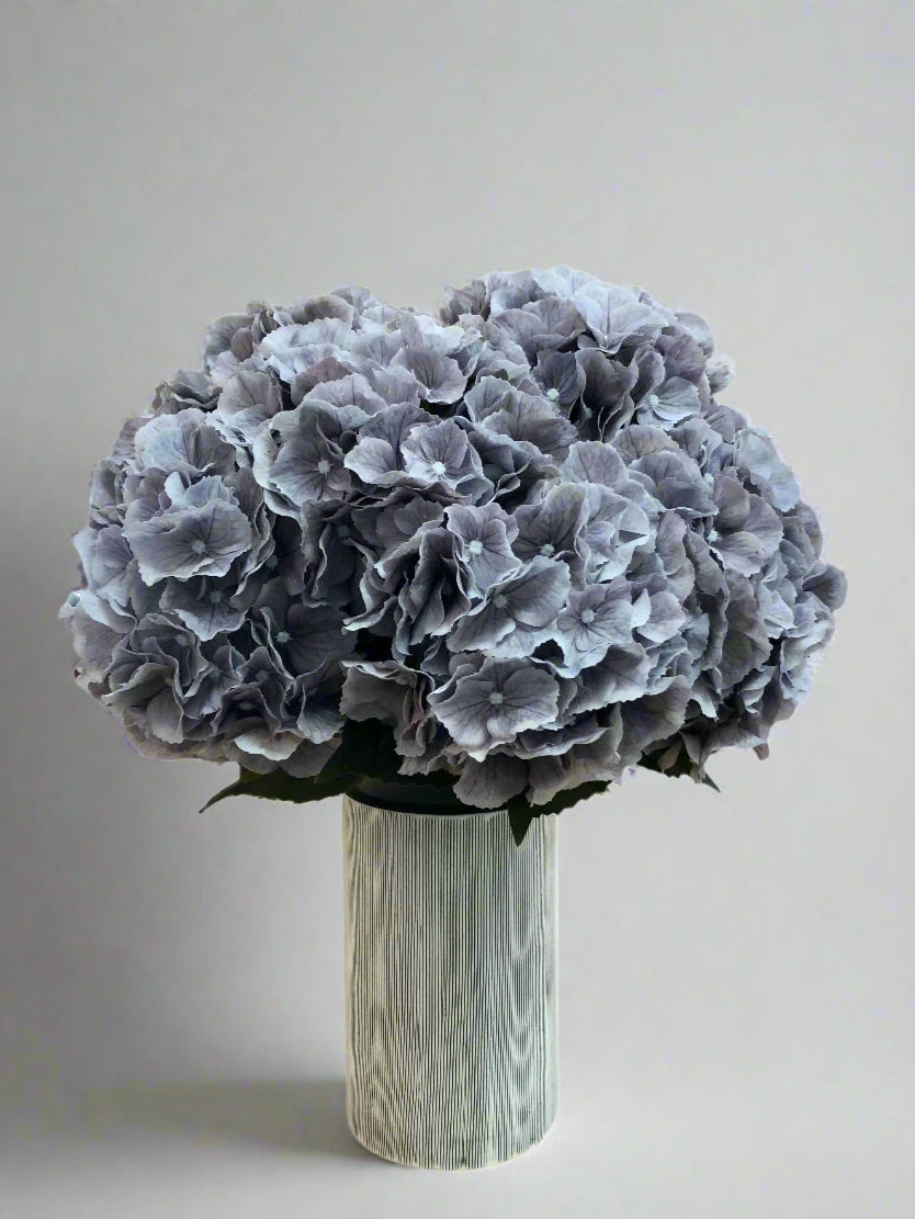 A high quality artificial grey Hydrangea flower arrangement displayed in a modern off white ceramic vase with charcoal pinstripe.