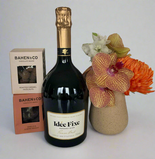 Bottle of Idee Fixe wine with 2 x box of Bahen & Co chocolate and a vase of flowers on a reflective surface.