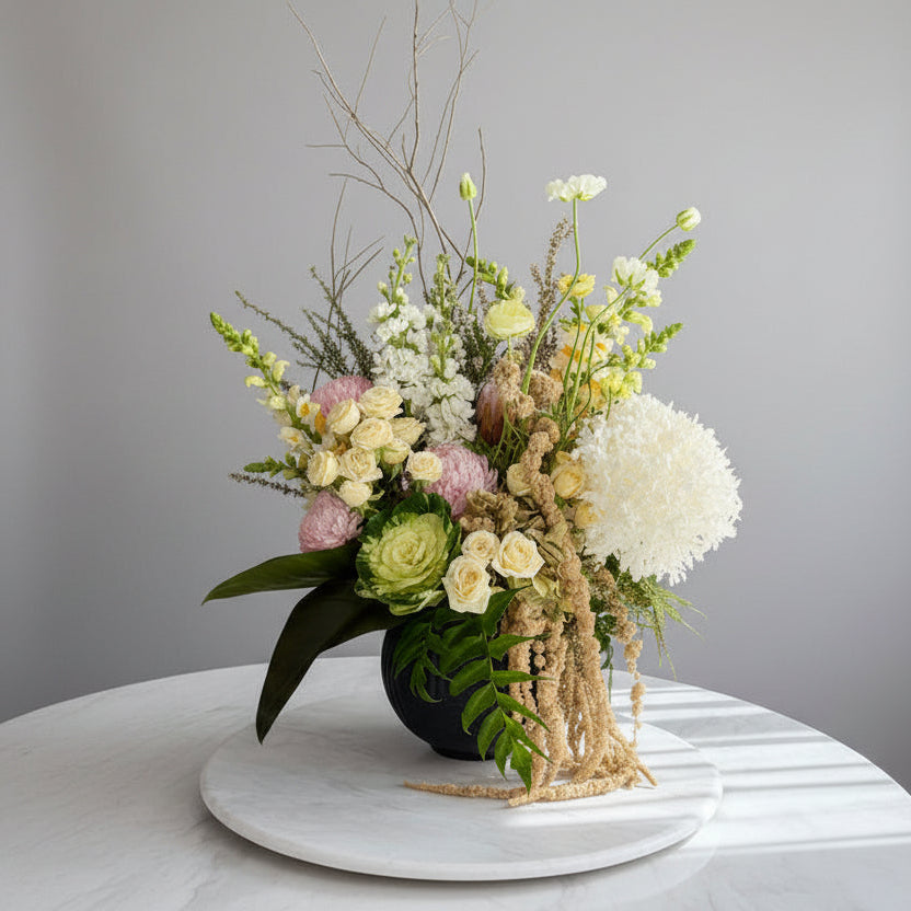 A large, lush floral arrangement featuring pale yellow roses, white hydrangea blooms, pink flowers, green foliage, and dried beige elements, displayed on a white marble-topped table in an indoor room.