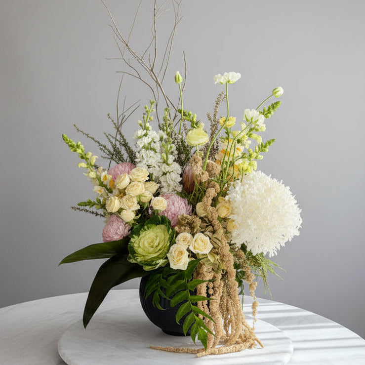 A large, lush floral arrangement featuring pale yellow roses, white hydrangea blooms, pink flowers, green foliage, and dried beige elements, displayed on a white marble-topped table in an indoor room.