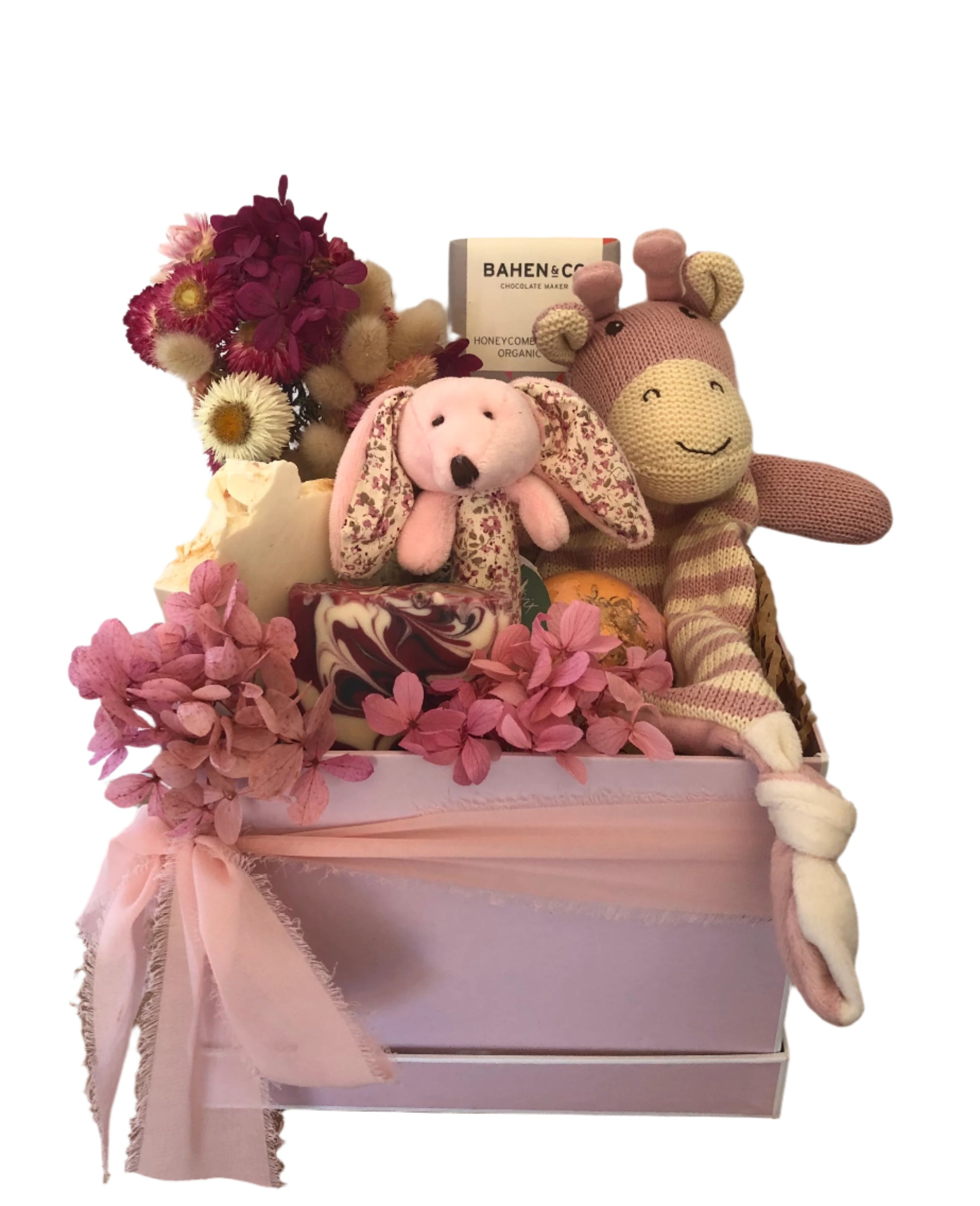 A pink box containing a variety of baby gifts including plush toys, a stick rattle, a comforter, organic milk chocolate, a bath bomb, and luxurious handmade soaps, adorned with dried flowers and a ribbon.