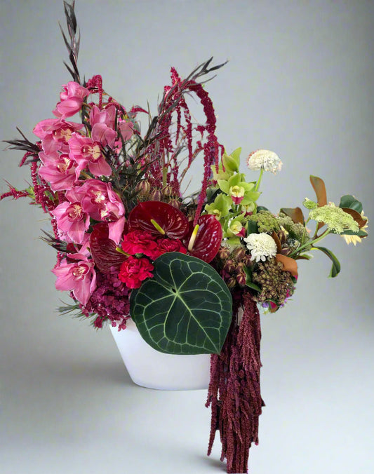 Colorful floral arrangement with pink orchids, red flowers, and green leaves in a white vase on a light gray background.