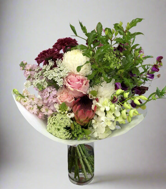 A bouquet of seasonal flowers arranged in a vase, with a mix of different flowers including roses and other foliage, wrapped in white paper.