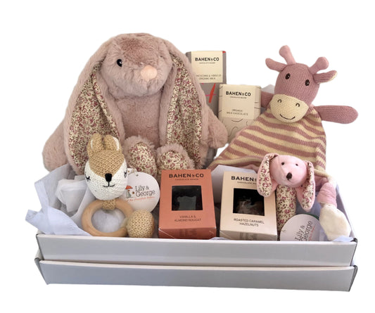A baby hamper gift set containing a plush bunny, a giraffe comforter, a teether, a stick rattle, and various chocolate candies. Presented in a white hamper box with ribbon.