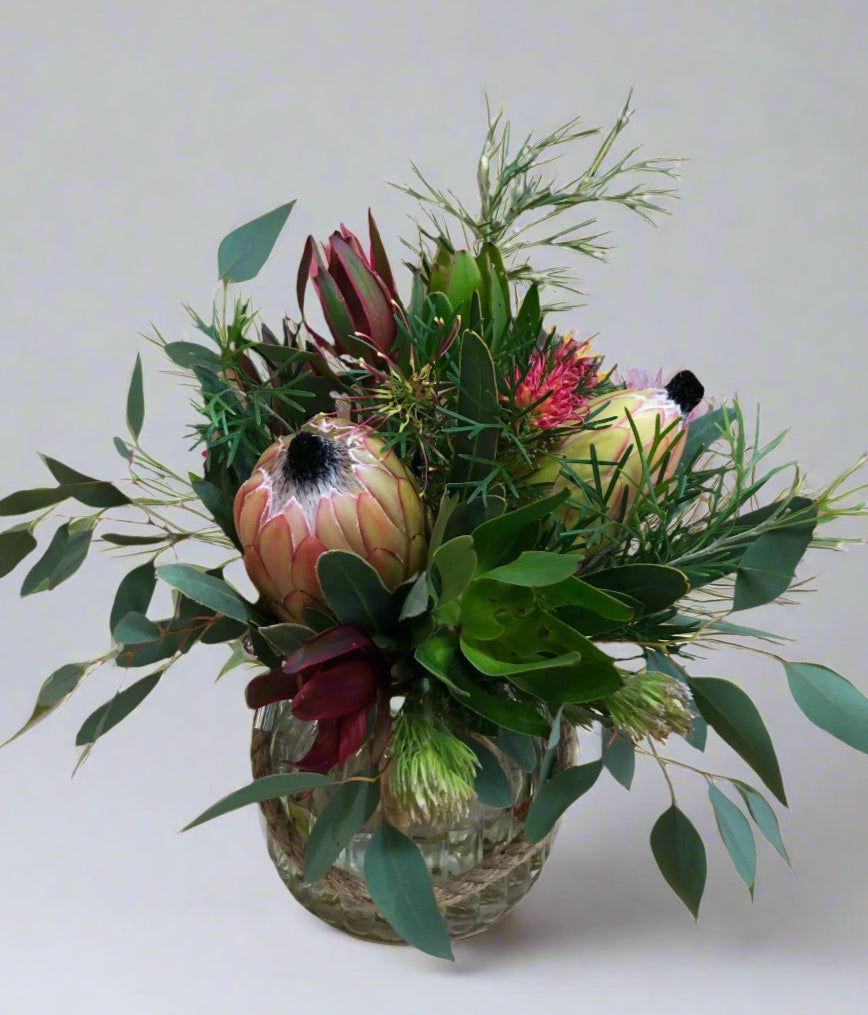 A floral arrangement featuring a selection of local natives and foliage in a glass vase with a rope handle.