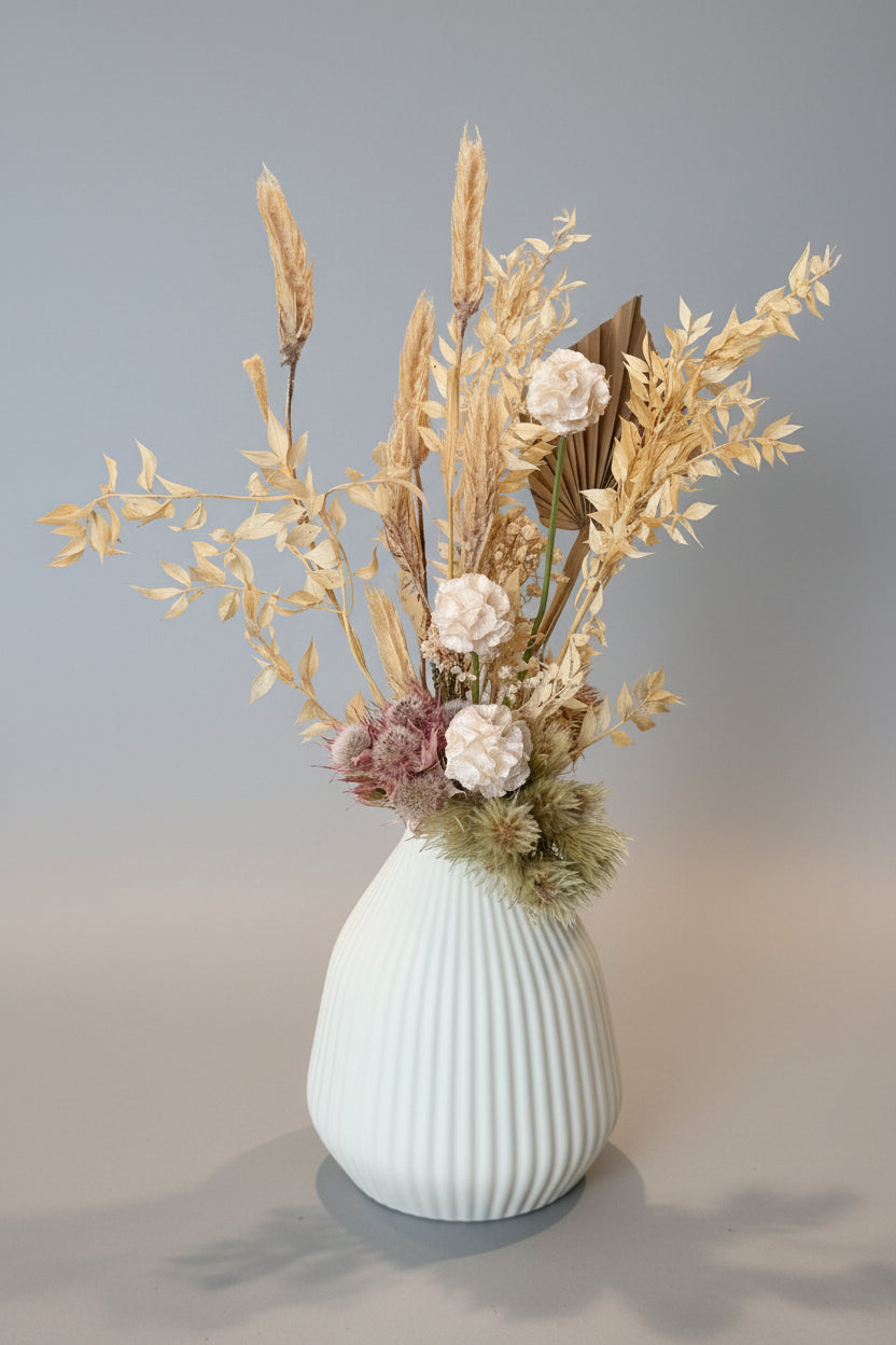 A dried floral arrangement featuring golden wheat stalks, beige foliage, and soft pink and cream flowers, presented in a ribbed white ceramic vase on a neutral indoor surface.