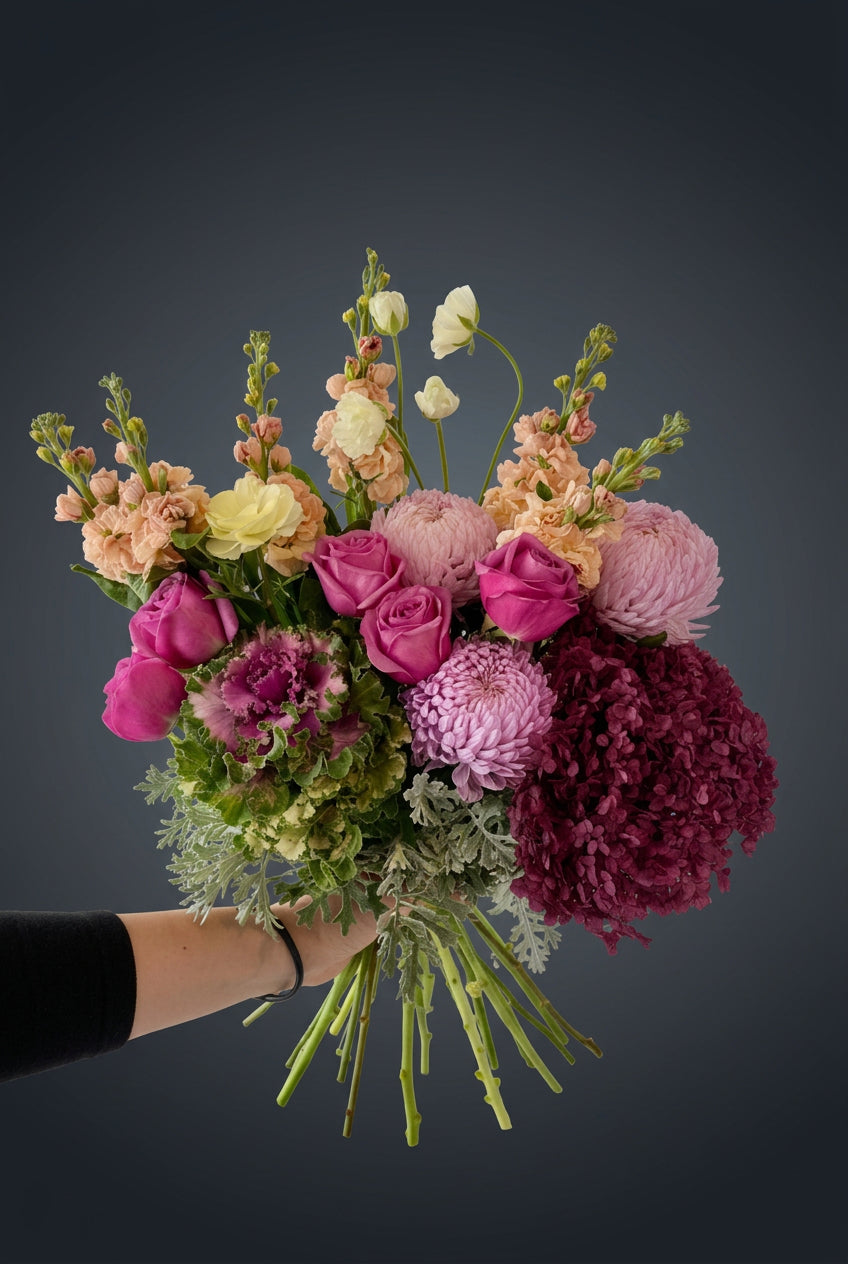 A vibrant, large floral bouquet held up by a person's hand in a workshop or storage room. The bouquet features a rich mix of pink, purple, peach, and green flowers, including roses, chrysanthemums, hydrangeas, ranunculus, stocks and kale.