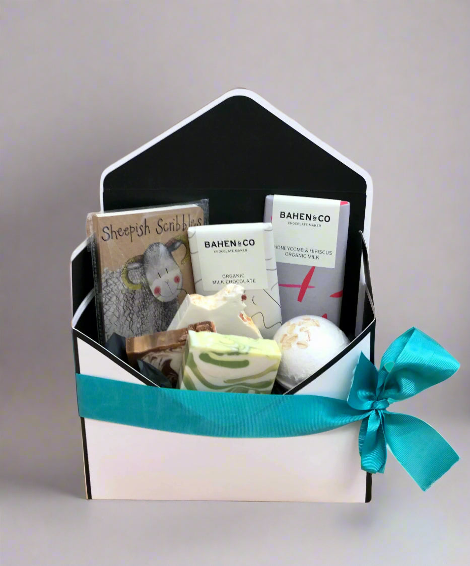 A gift basket hamper presented in an envelope box with a variety of items including a mini notebook, bath bomb, handmade soaps, and chocolates, tied with a blue ribbon.