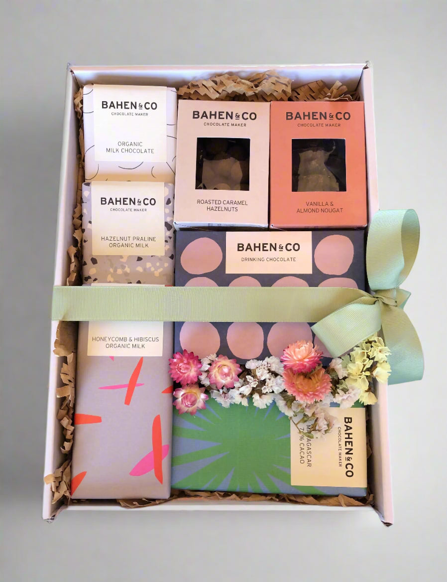 A gift hamper box containing various chocolate items from Bahen & Co, including drinking chocolate, roasted caramel hazelnuts, vanilla & almond nougat, and milk chocolate pieces, decorated with dried flowers and a green ribbon.