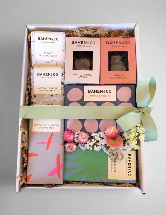 A gift hamper box containing various chocolate items from Bahen & Co, including drinking chocolate, roasted caramel hazelnuts, vanilla & almond nougat, and milk chocolate pieces, decorated with dried flowers and a green ribbon.