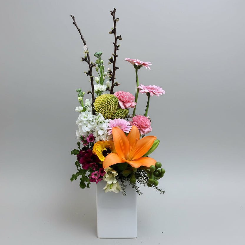Colorful flower arrangement with an orange lily, pink and white flowers, and greenery on a brown background.