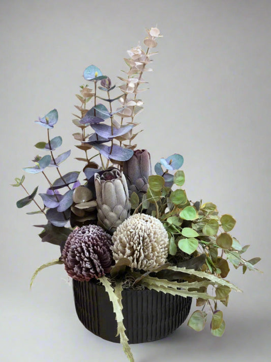 Artificial flower arrangement including Banksias, Proteas, Gum, and foliage in a recycled paper vessel with reindeer moss.