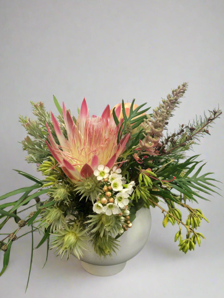 Floral arrangement with a prominent pink protea in a white vase on a plain background