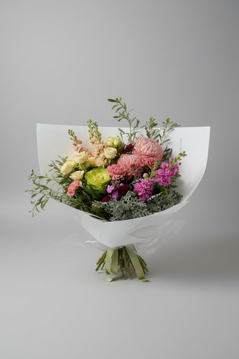 A wrapped flower bouquet with pink, purple, green, and cream blossoms, wrapped in white paper, set against a solid gray background.
