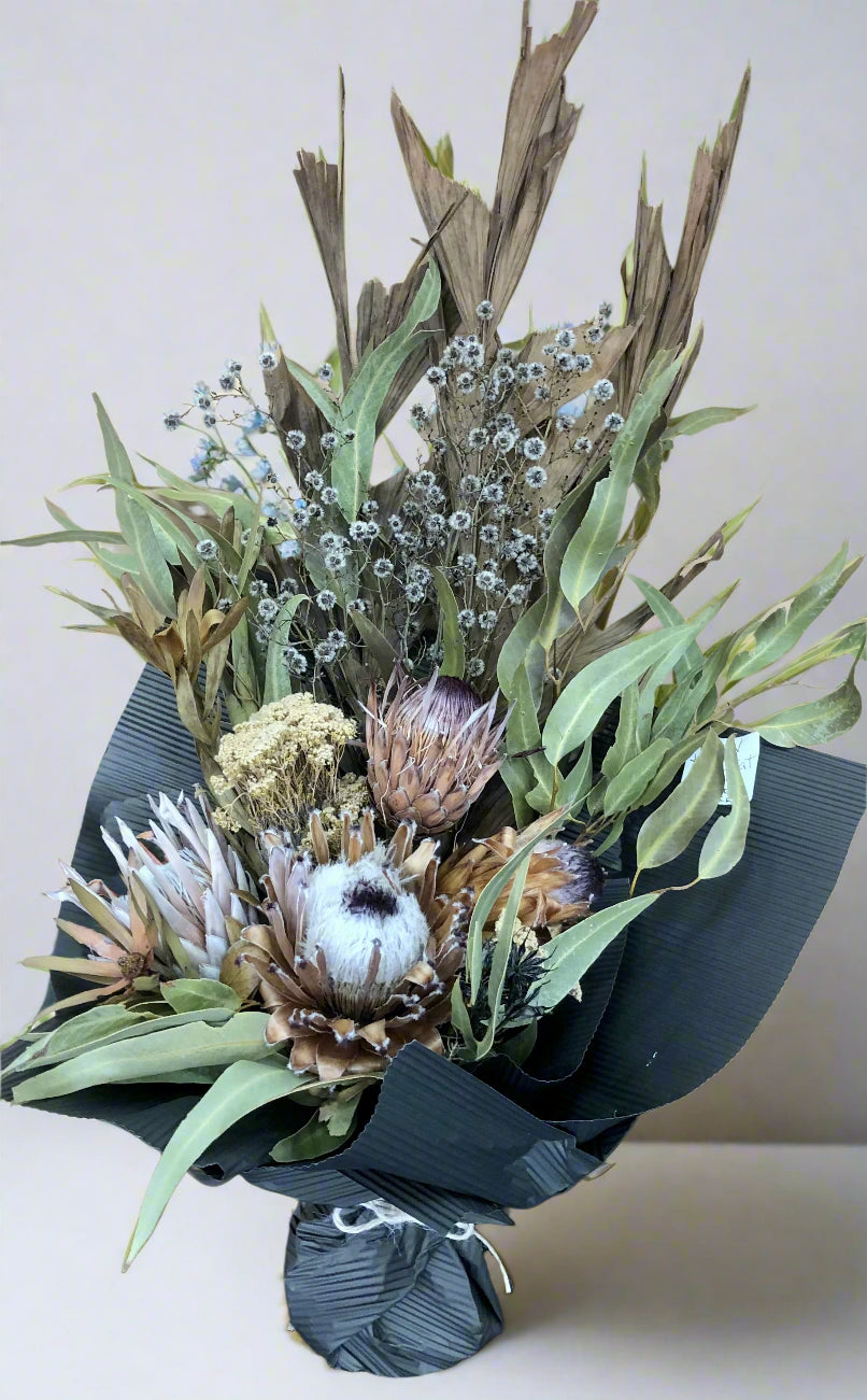 A dried native bouquet featuring a variety of flowers including Proteas, Leucadendron, and Stirlingia, with Fish Tail Palm and Gum leaves, arranged in a naturalistic style on a white background.