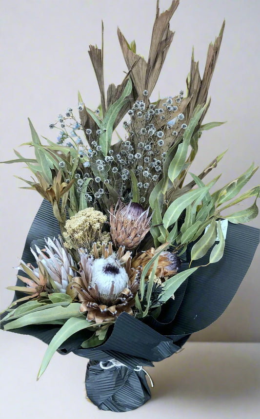 A dried native bouquet featuring a variety of flowers including Proteas, Leucadendron, and Stirlingia, with Fish Tail Palm and Gum leaves, arranged in a naturalistic style on a white background.