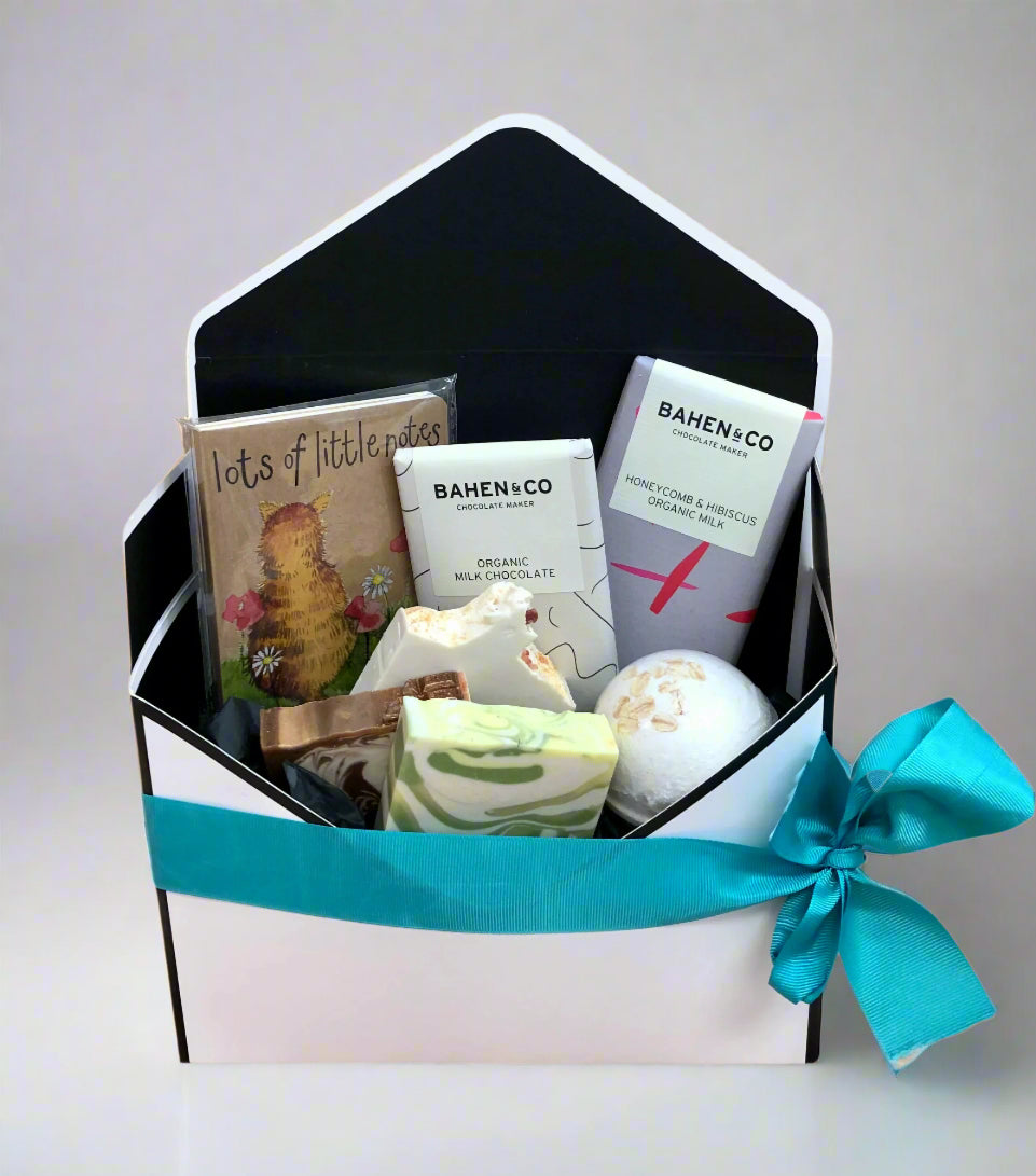 A gift hamper box with various items including a mini notebook, chocolates, bath bombs, and handmade soaps, tied with a blue ribbon.