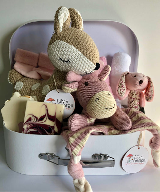 A baby hamper gift set containing plush toys, a comforter, a rattle, socks, wash cloths, and handmade soaps, presented in a suitcase-style box.