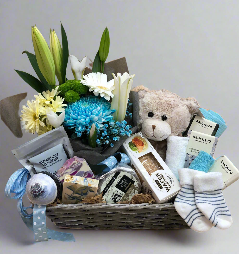 A wicker basket gift hamper with a bouquet of flowers, plush teddy bear, baby wash cloth, bath bomb, chocolates, and other baby items.