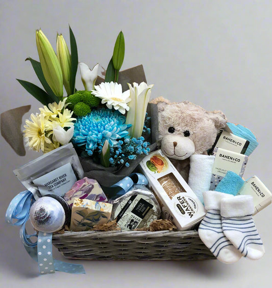 A wicker basket gift hamper with a bouquet of flowers, plush teddy bear, baby wash cloth, bath bomb, chocolates, and other baby items.