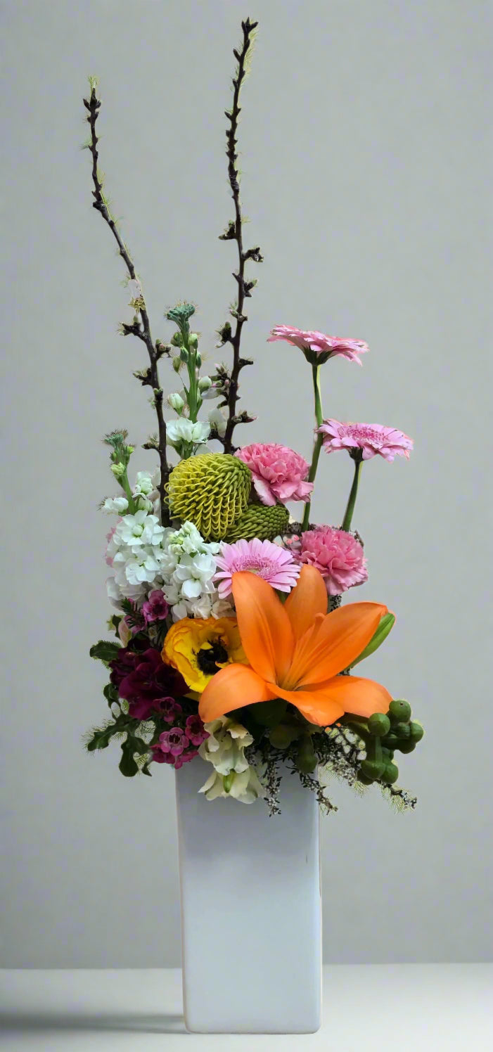 A floral arrangement featuring a variety of flowers such as lilies and gerberas, with green foliage and branches, in a white ceramic vase.