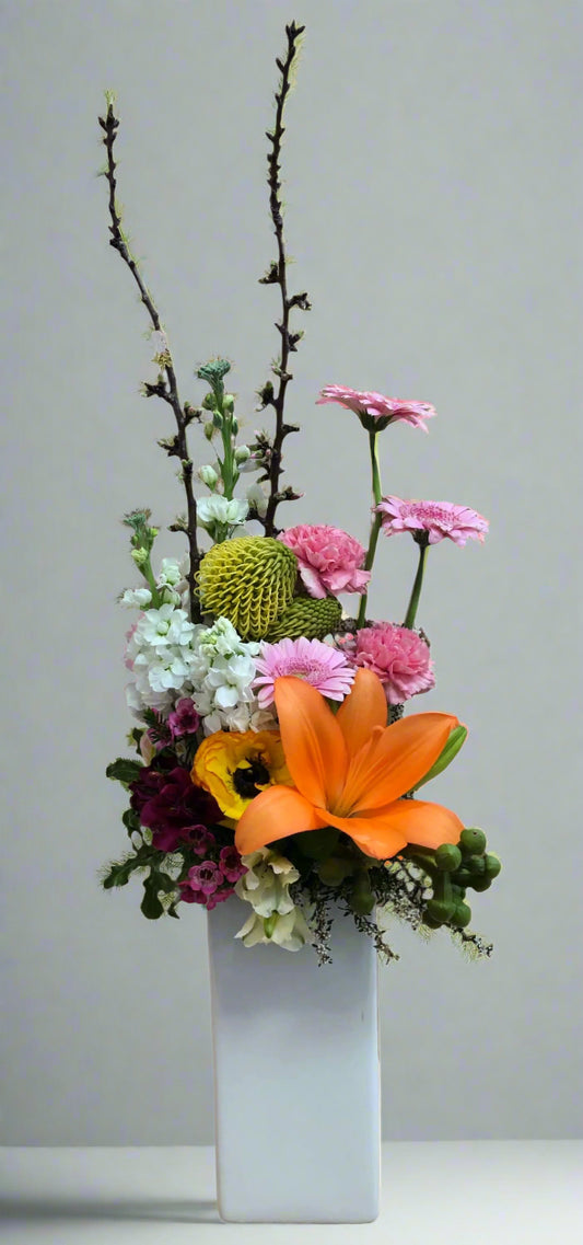 A floral arrangement featuring a variety of flowers such as lilies and gerberas, with green foliage and branches, in a white ceramic vase.