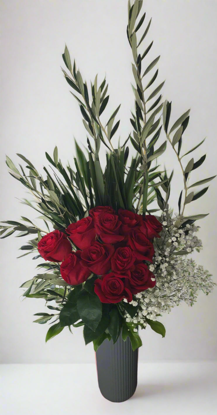 Vase with red roses and greenery on a wooden surface against a white wall