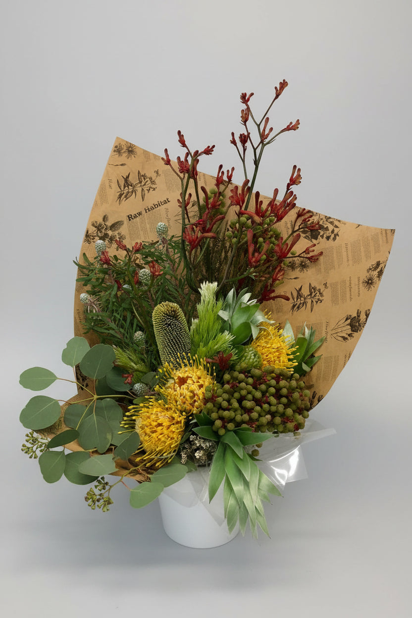 A mixed floral arrangement featuring yellow protea-like flowers, green and red berries, eucalyptus leaves, and reddish-brown stems, wrapped in brown paper with botanical illustrations, set against a white textured wall.