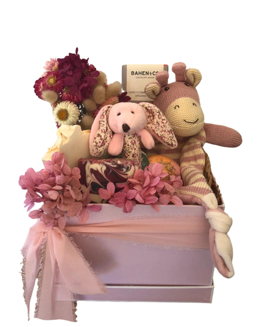 A pink box containing a variety of baby gifts including plush toys, a stick rattle, a comforter, organic milk chocolate, a bath bomb, and luxurious handmade soaps, adorned with dried flowers and a ribbon.