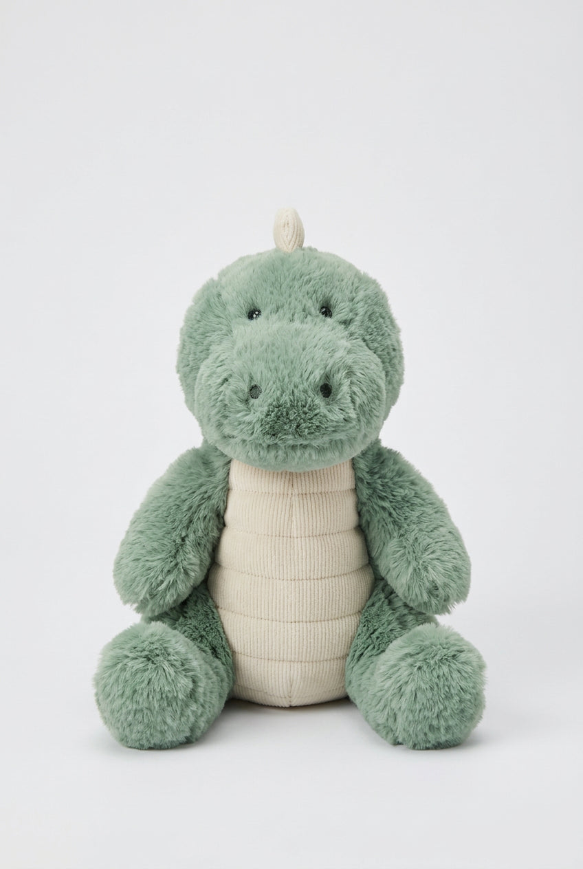 A plush dinosaur toy in sage green with a cream-colored belly, sitting on a round wooden surface against a white background.