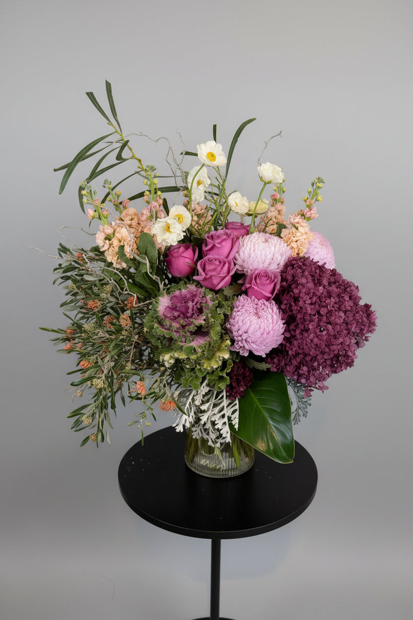 A vibrant floral arrangement in a black round vase, featuring pink and purple roses, light pink peonies, burgundy hydrangeas, and green foliage, set against a neutral indoor background.