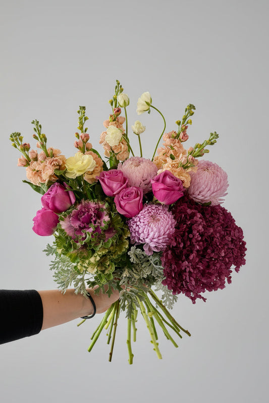 A vibrant, large floral bouquet held up by a person's hand in a workshop or storage room. The bouquet features a rich mix of pink, purple, peach, and green flowers, including roses, chrysanthemums, hydrangeas, ranunculus, stocks and kale.