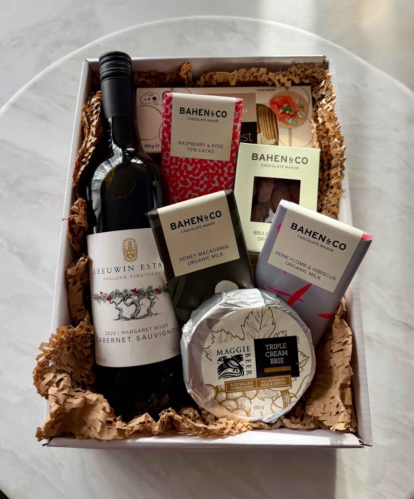 A curated gourmet gift box filled with food items, including a bottle of Lievin Estate Cabernet Sauvignon, a round cheese wheel labeled Maggie's, and multiple packages from Bahen+Co, arranged in brown crinkle paper inside a white box on a light marble surface.