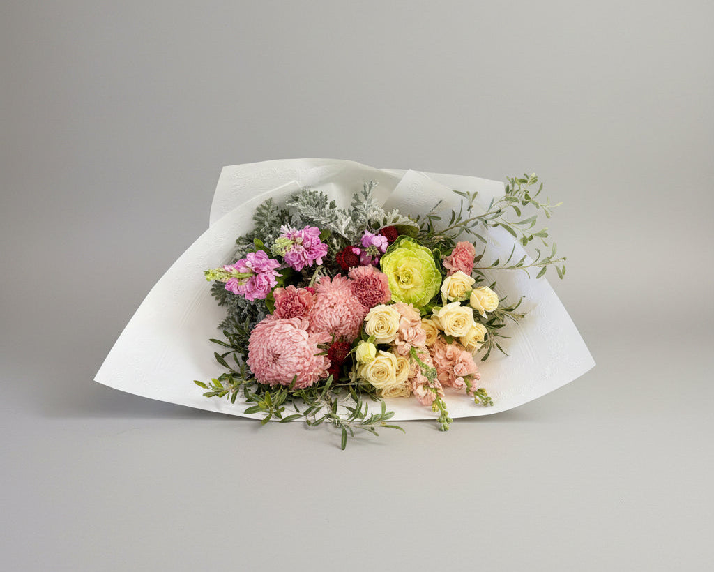A floral bouquet wrapped in white paper, featuring pink and cream flowers with green foliage, placed on a gray concrete floor.