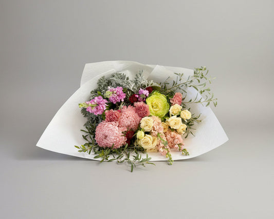 A floral bouquet wrapped in white paper, featuring pink and cream flowers with green foliage, placed on a gray concrete floor.