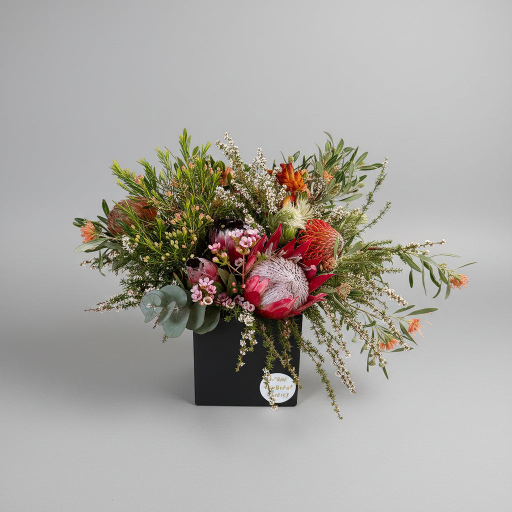 A vibrant floral arrangement featuring protea flowers, pink blossoms, and green foliage in a square black container, placed on a gray concrete floor in an indoor workspace.