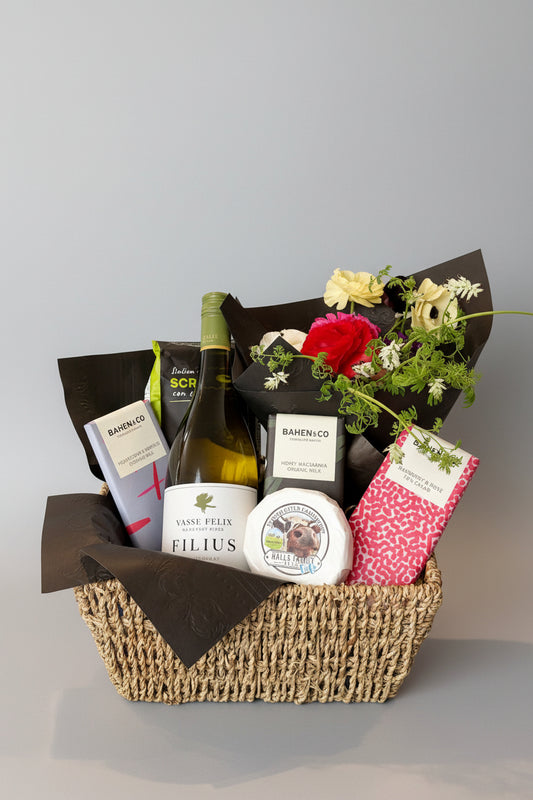 A curated gift basket in a woven straw basket, filled with gourmet items including a bottle of Vasse Felix Filius white wine, a wheel of cheese, crackers ,Bahen & co Chocolates, and a bouquet of fresh flowers wrapped in black paper