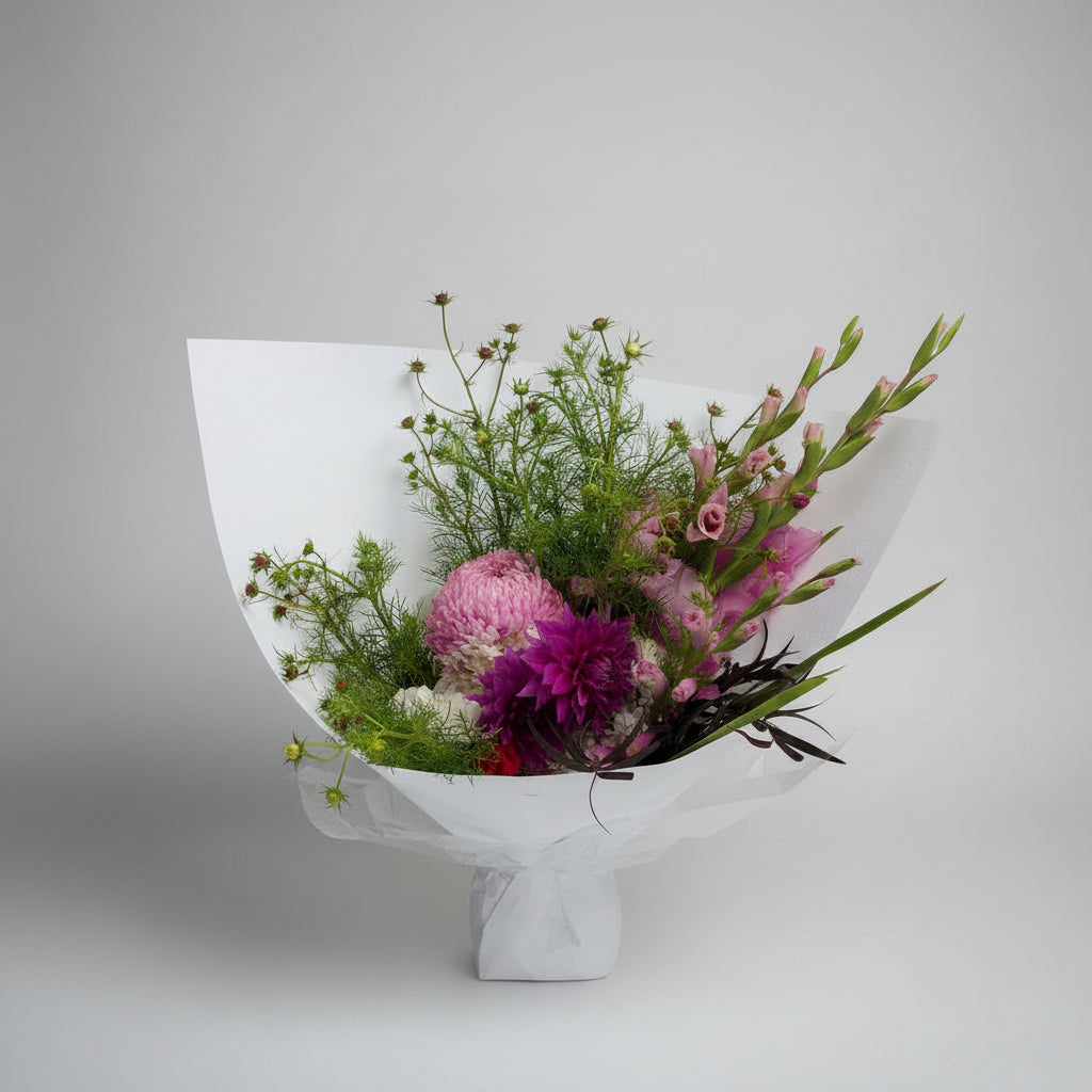 A mixed flower bouquet wrapped in white paper, featuring pink and purple flowers with green foliage, photographed indoors against a light curtain background.