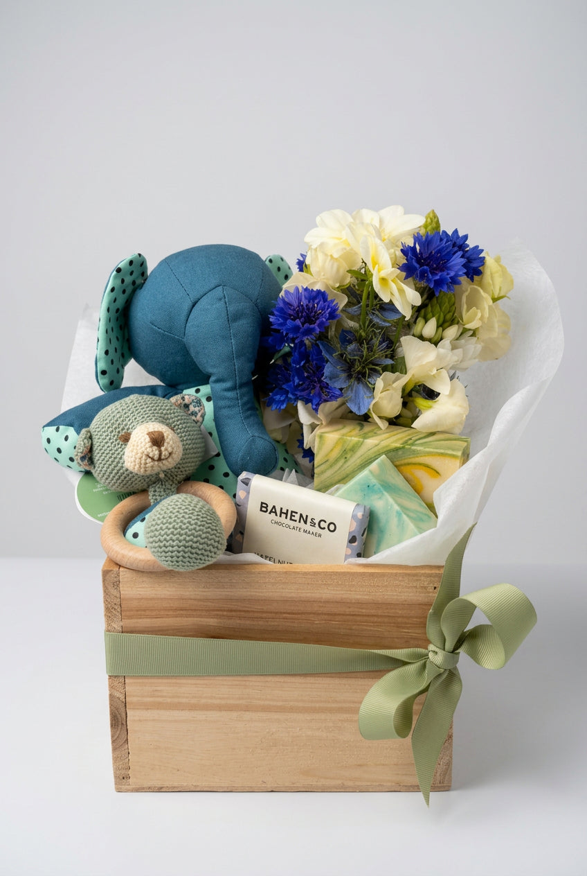 A baby gift basket arranged in a wooden box with a sage green ribbon, containing a blue plush elephant toy, a knitted bear teether, a bar of Bahen & Co soap, and a bouquet of white and blue flowers wrapped in white tissue paper.