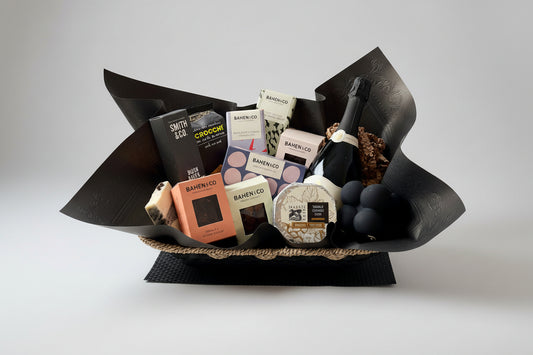 A gourmet gift basket filled with assorted cheeses, crackers, chocolate, and a bottle of champagne, wrapped in black paper and arranged on a white plate.