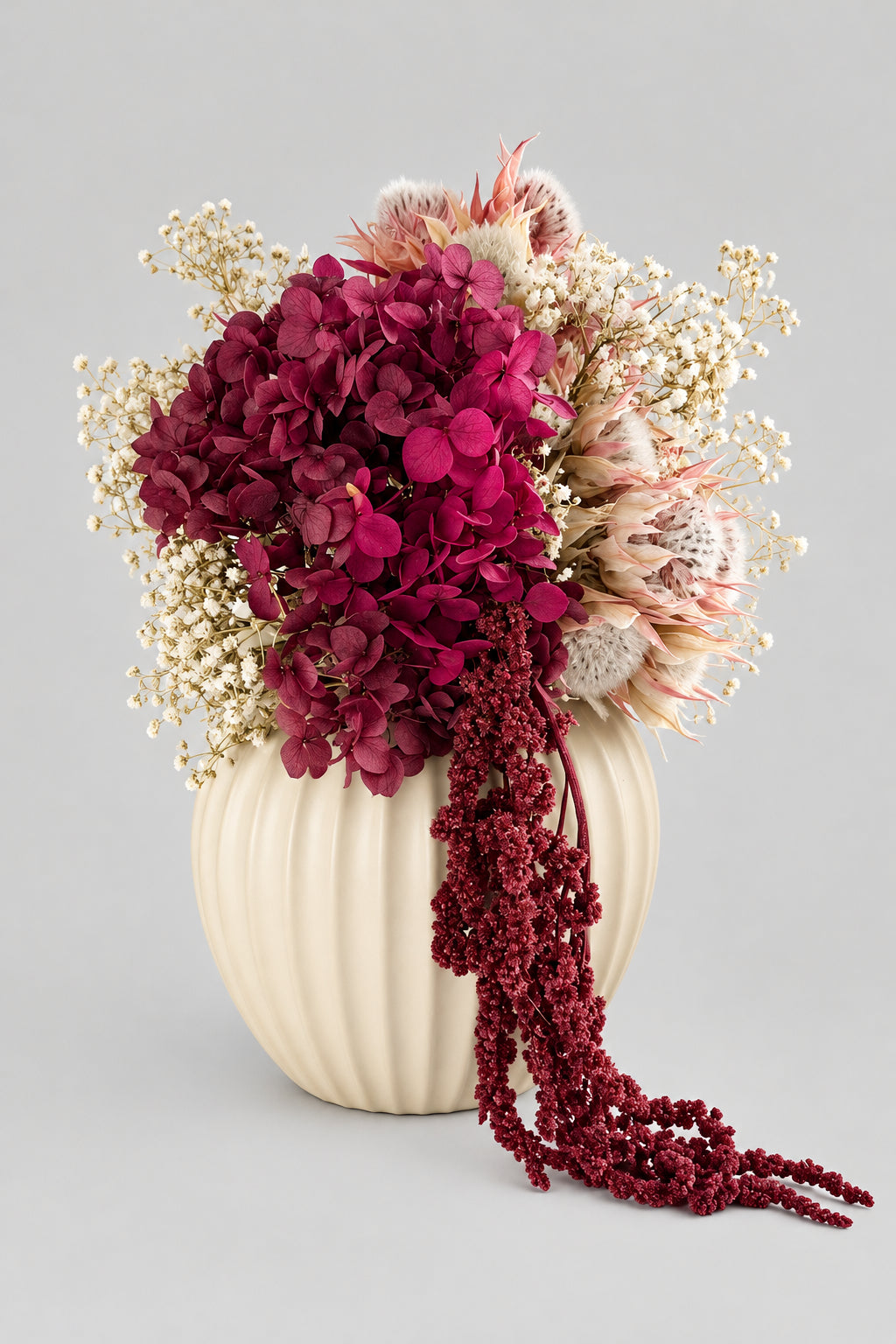 A dried floral arrangement in a ribbed cream ceramic vase, featuring deep magenta hydrangeas, beige baby's breath, and a long cascading burgundy beaded strand, displayed on a white surface.