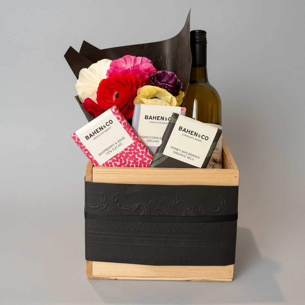 A gift basket arrangement in a wooden crate with a black ribbon, featuring a bottle, a bouquet of a flowers, and BAHEN+CO chocolate bars on a concrete floor.
