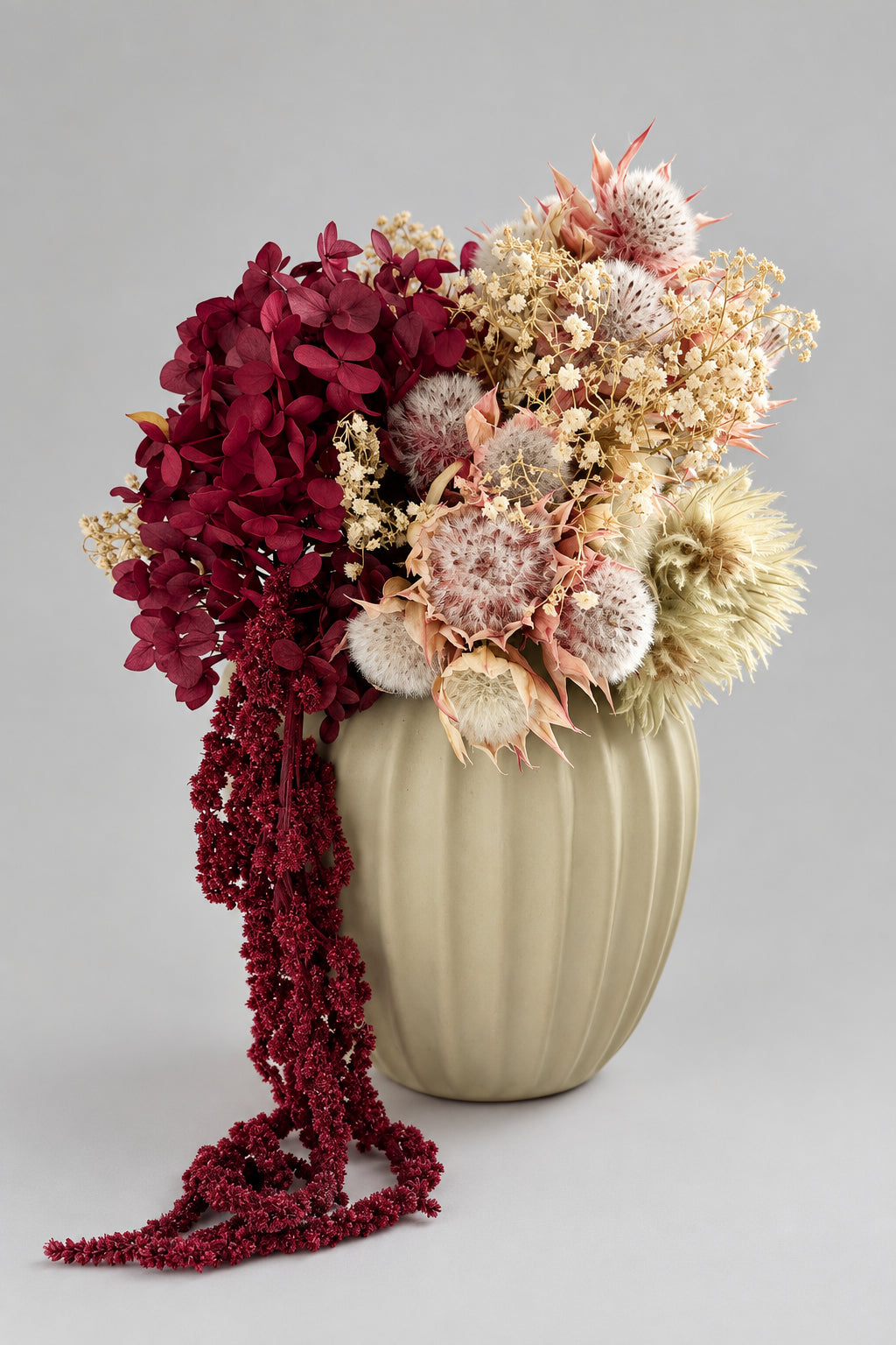 A dried flower arrangement in a ribbed cream-colored vase, set on a white surface with a blurred background showing a craft or floral workspace.